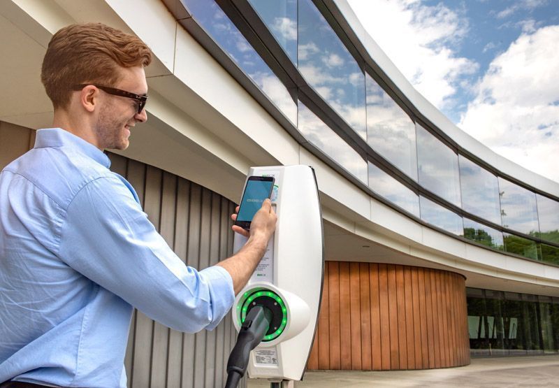 A young professional wearing sunglasses and an unironed shirt standing in front of a workplace next to an EVBox Business Line EV charging station, while opening the EVBox app on his smartphone.