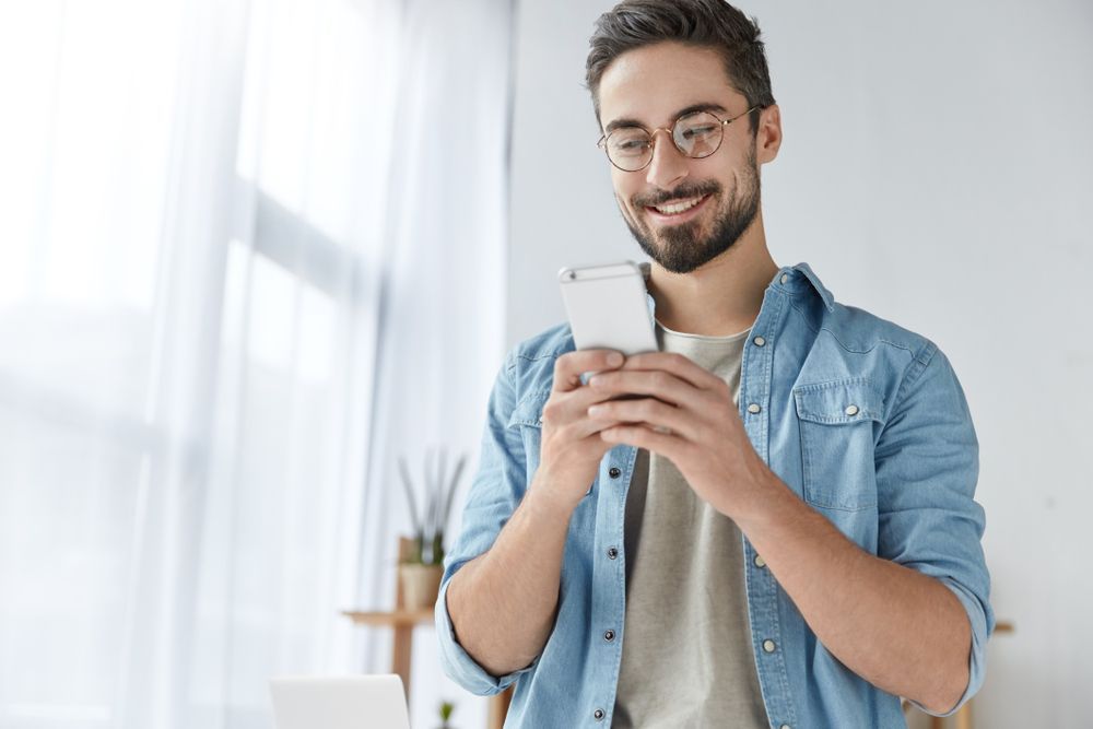 A bearded man wearing glasses and a casual outfit stands in his living room smilingly holding his smartphone while checking out the valuable insights he has gained from his EV charging app.