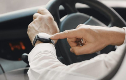 A zoomed in shot of 2 hands of a man behind the wheel of a parked car who's pointing at his watch.