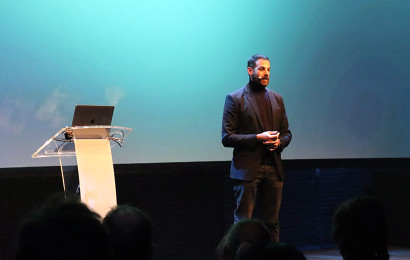 A man giving a speech on stage with a podium next to him against a dark blue and teal backdrop.