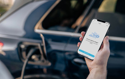 A hand holding a phone showing an EV charging app next to a charging EV