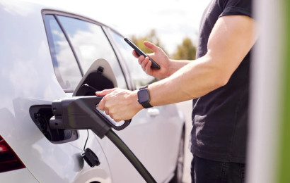 A man inserting a cable from the fast charger to his electric car while holding his smartphone in the other hand.