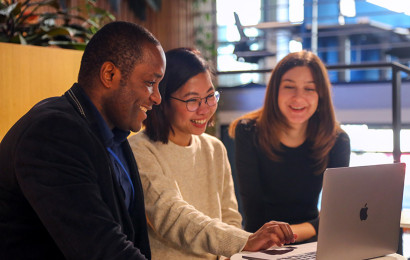 A man and two women looking at a laptop in the middle of them, they are all smiling at the laptop.